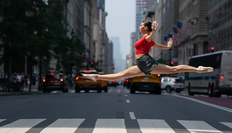 Dancer performing a grand jeté across a city street crosswalk with yellow taxis and urban buildings in background.