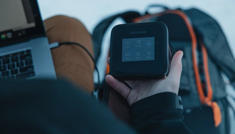 Person holding a portable WiFi hotspot device with a small LCD screen while sitting near a laptop and backpack.