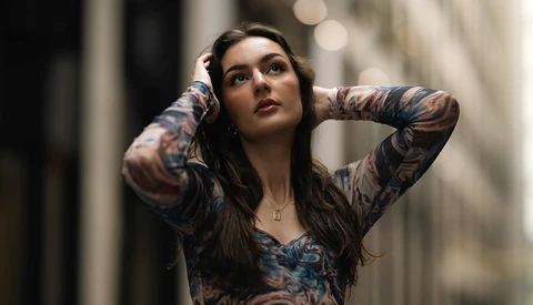 Woman with full-sleeve tattoos looking upward with hands behind head in urban alley.
