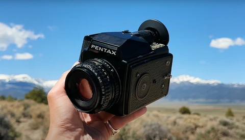 A Pentax 67 medium format camera held in someone's hand against a mountain landscape backdrop.