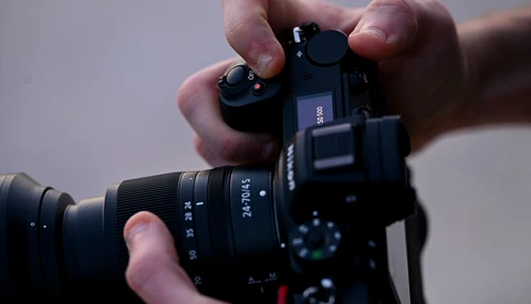 Hands holding a mirrorless camera with attached lens, viewed from above against a neutral background.