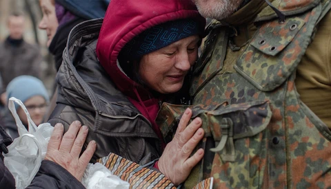 Woman in burgundy winter hat smiling while embraced by two people in military-patterned uniforms.