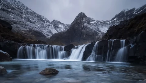 Tiered waterfalls flowing into a blue pool beneath snow-capped mountains at twilight.