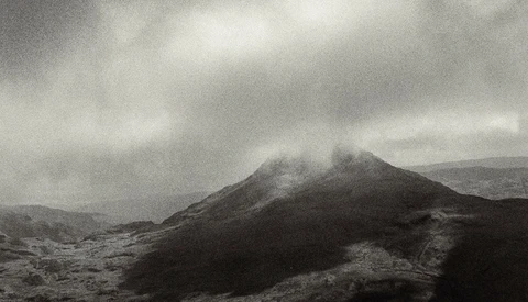 Volcanic cone shrouded in mist under overcast sky with barren lava fields in foreground.