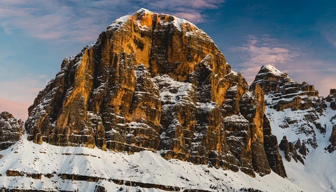 Snow-covered rocky mountain peak with golden-orange columnar formations lit by warm sunlight against blue sky.