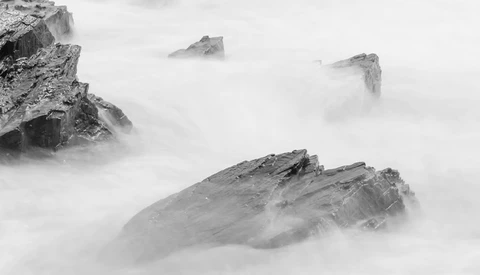 Rocky outcrops partially submerged in thick fog above turbulent ocean water.