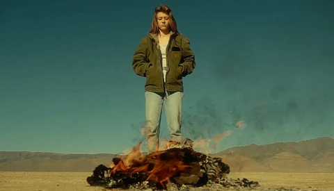 Person standing on burning debris in desert landscape under teal sky.