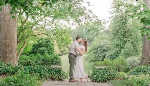 Couple kissing on a stone pathway lined with tall trees and lush green foliage.