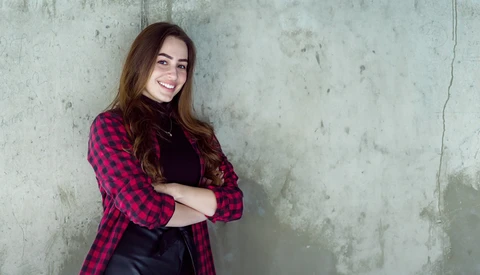 Woman in red plaid shirt leaning against weathered concrete wall with arms crossed.
