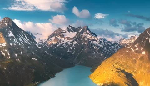Alpine landscape with turquoise glacial lake nestled between snow-capped mountain peaks under partly cloudy sky.