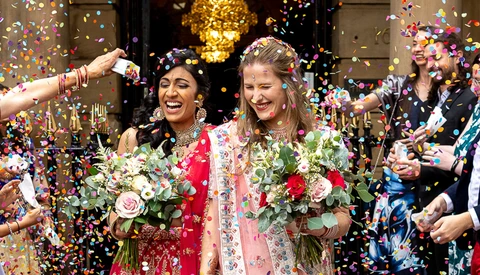 Two people in traditional wedding attire smiling amid falling colorful confetti.