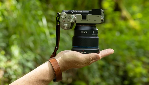 Hand holding a compact mirrorless camera with attached lens against a blurred green outdoor background.