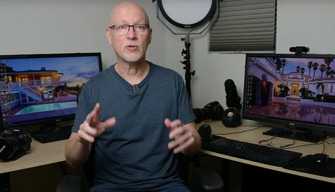 Man in glasses and blue shirt gesturing while speaking in a studio with multiple monitors displaying architectural renderings.