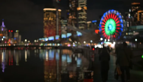 Illuminated Ferris wheel reflected in still water at night with blurred city skyline behind it.