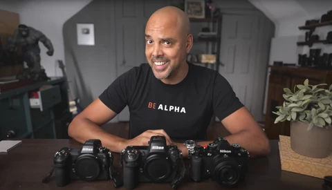 Man in black 'BE ALPHA' shirt displays three professional DSLR cameras on a desk.