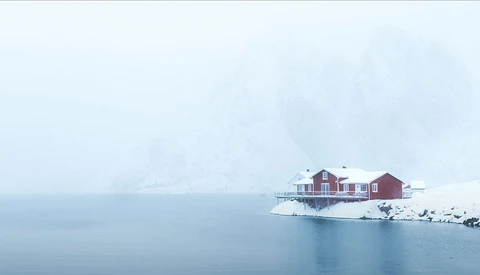 Red houses on a snowy peninsula surrounded by still, misty water in winter conditions.