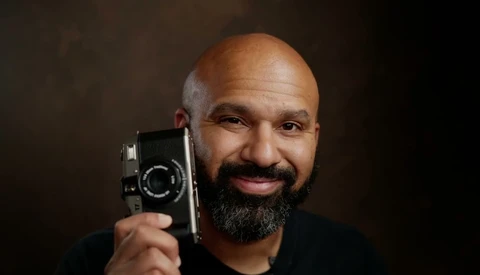 Bearded man holding a vintage film camera up to his face against a dark backdrop.
