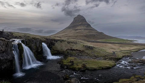 Cone-shaped mountain with cascading waterfalls in foreground under dramatic cloudy sky.