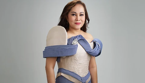 Woman in cream-colored off-shoulder dress with blue striped fabric draped around shoulders against neutral background.