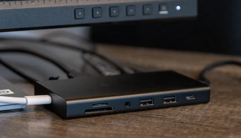 Docking station with multiple ports connected to a laptop, sitting on a wooden desk next to a monitor.