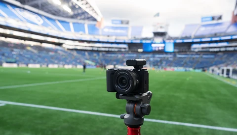 Professional camera mounted on tripod positioned at field level inside a large stadium with blue seating.