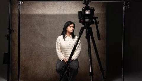 Woman in cream knit sweater seated in studio with camera mounted on tripod and professional lighting setup.