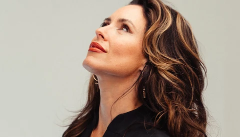 Woman with wavy brunette hair and red lipstick gazing upward against neutral background.