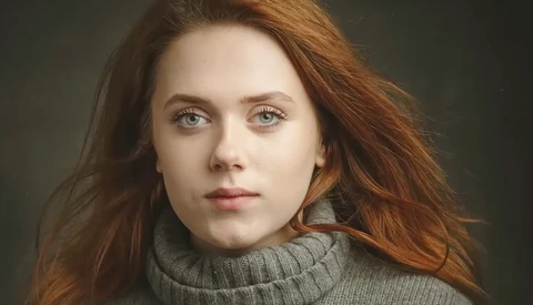 Woman with long reddish-brown hair wearing a gray turtleneck against a dark green background.