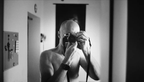 Bald man holding vintage camera up to his face in a mirror self-portrait.