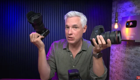 Man holding two camera lenses while speaking in a studio with purple and blue lighting.