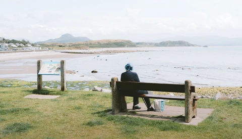 Person sitting on wooden bench overlooking a coastal beach with mountains in the distance.