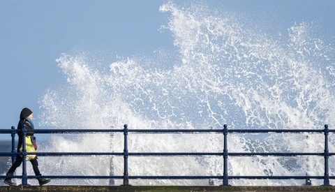 Person in yellow safety vest standing at railing as large wave crashes against barrier.