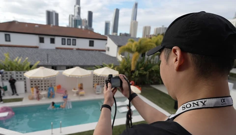 Photographer holding mirrorless camera with Sony strap, shooting poolside scene with city skyline beyond.