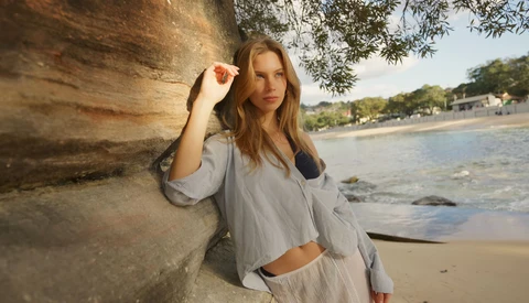 Woman in light blue shirt leaning against weathered wooden post on beach at golden hour.