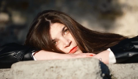 Woman with long dark hair and red lipstick resting her head on a concrete surface, looking at the camera.