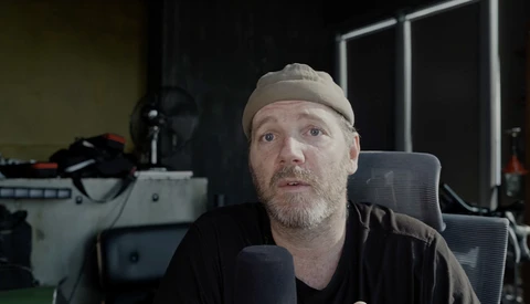 Man wearing a beanie and dark shirt speaking into a microphone in a studio setting.