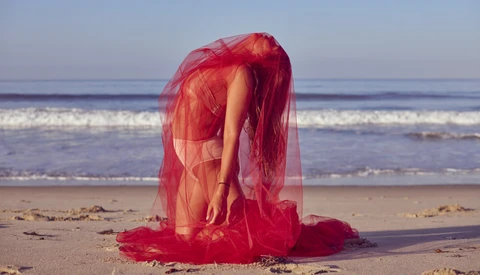 Person wrapped in sheer red fabric on a beach, bent forward with arms extended toward the sand.
