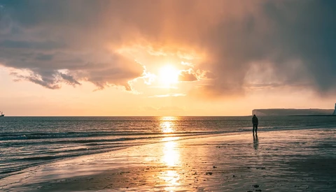 Surfer standing in shallow water during golden hour sunset with dramatic clouds and bright sun breaking through.