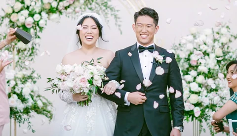 Bride and groom smiling at wedding ceremony with floral backdrop and petal confetti.