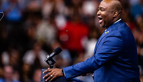 Man in blue blazer speaking at microphone before a crowd.