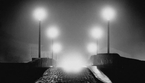 Industrial street lined with street lamps receding into fog, rendered in high-contrast black and white.
