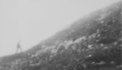 Grainy black and white photograph of a hillside with scattered rocks and sparse vegetation under overcast sky.