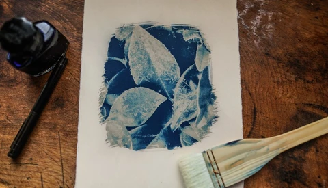 Cyanotype print with blue botanical leaf shapes on white paper, surrounded by art materials on wood surface.