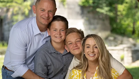 Family of four smiling outdoors in casual clothing with green foliage background.