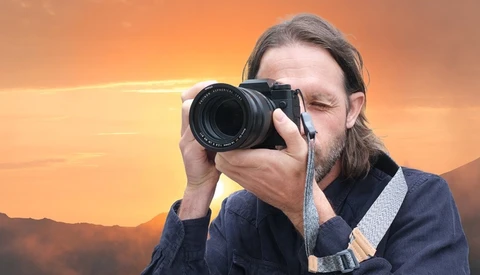 Photographer with long hair and beard holding a telephoto lens camera to his eye against a golden sunset sky.