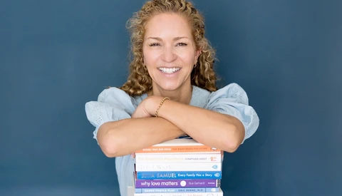 Woman with curly blonde hair smiling at camera while leaning on a stack of colorful books against blue background.