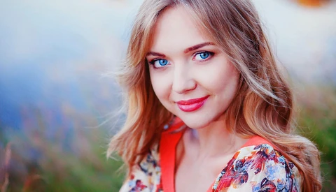 Woman with reddish-brown wavy hair and bright blue eyes, wearing red lipstick and a colorful patterned garment outdoors.