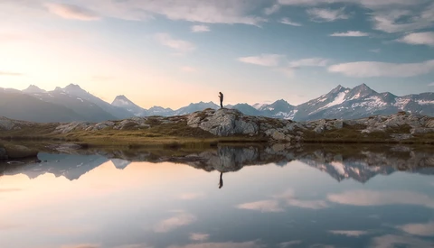 Solitary hiker standing on a rocky outcrop, reflected in still water with snow-capped mountains in the background.