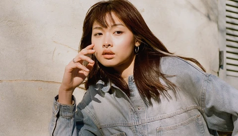 Woman in denim jacket with hand near face, shot against textured neutral wall.