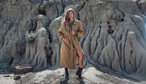 Woman in tan trench coat and black boots standing against dramatic eroded clay badlands formation.